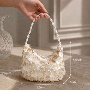 May include: A small handbag covered in iridescent white circular shell-like discs with a handle made of white beads. The bag measures 6.7 inches in width, 5.5 inches in height, and 1.2 inches in depth, featuring a top zipper closure.