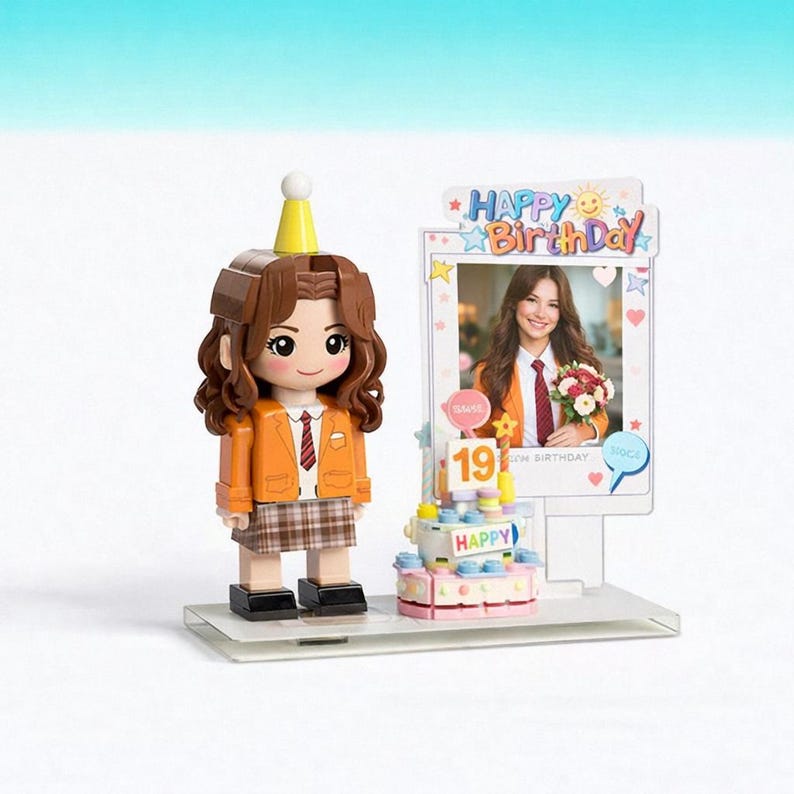 May include: A block-style figurine with brown hair wearing a yellow party hat, orange blazer, and plaid skirt, standing next to a birthday cake and a photo frame displaying a person holding flowers with the text Happy Birthday and the number 19.