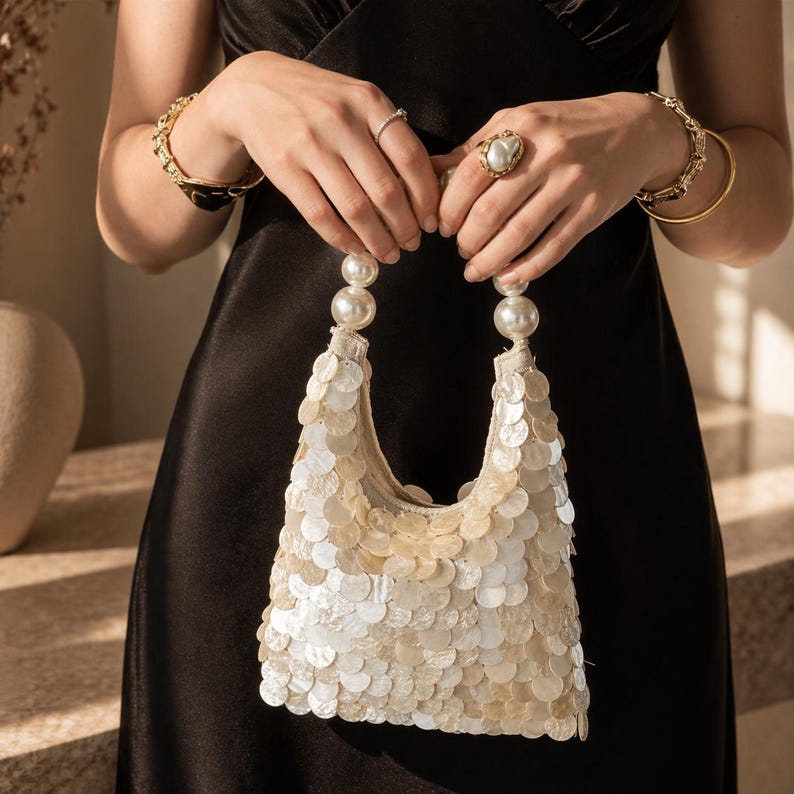 May include: A white sequin handbag with pearl handles is held by a person wearing gold bracelets and a ring. The bag is covered in small, round, iridescent sequins. The handles are made of large, white pearls.