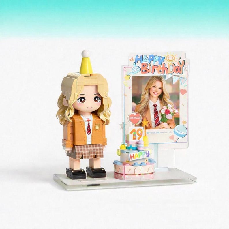 May include: A buildable plastic figure with blonde hair wearing a yellow party hat, tan blazer, and plaid skirt. Beside it sits a small tiered birthday cake with the number 19 and a vertical frame displaying a photo with the text Happy Birthday.