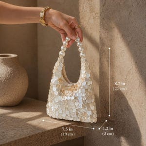 May include: Ivory sequin handbag with pearl beaded handles. The bag is 7.5 inches (19 cm) wide, 8.7 inches (22 cm) tall, and 1.2 inches (3 cm) deep. The bag is held by a person.