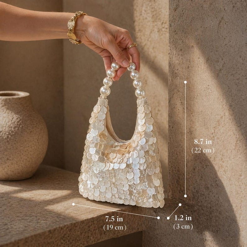 May include: Ivory sequin handbag with pearl beaded handles. The bag is 7.5 inches (19 cm) wide, 8.7 inches (22 cm) tall, and 1.2 inches (3 cm) deep. The bag is held by a person.