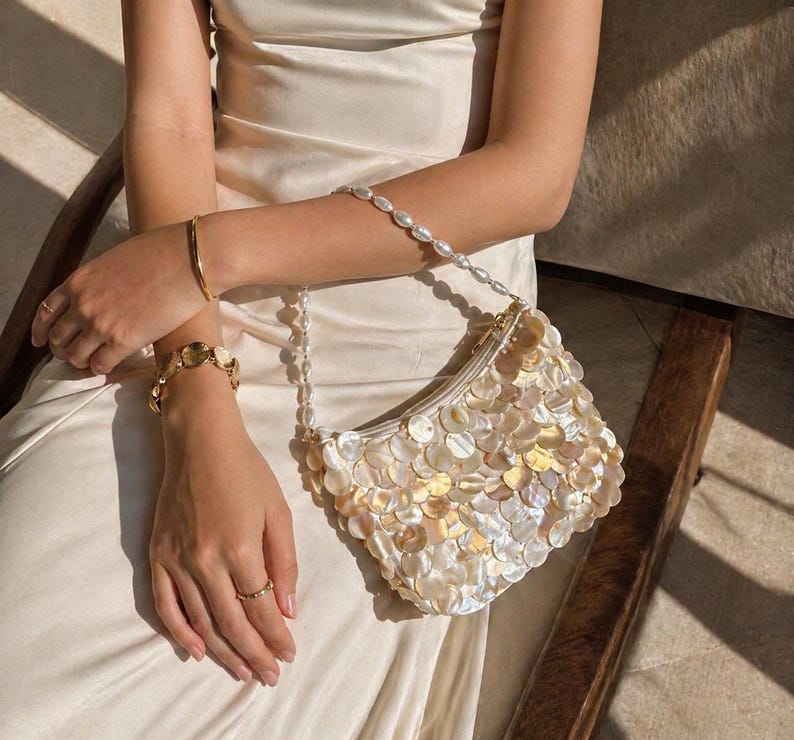 May include: A small handbag covered in iridescent, circular shell-like discs in shades of cream and pale gold. The bag features a shoulder strap made of white oval pearls. The handbag is held against a smooth, cream-colored satin fabric garment.