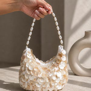 May include: A crescent-shaped handbag covered in overlapping iridescent mother-of-pearl discs. The bag features a top handle made of a single strand of white oval pearls and a gold-toned zipper closure at the top opening of the white fabric interior.