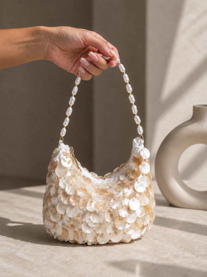 May include: A crescent-shaped handbag covered in overlapping iridescent mother-of-pearl discs. The bag features a top handle made of a single strand of white oval pearls and a gold-toned zipper closure at the top opening of the white fabric interior.