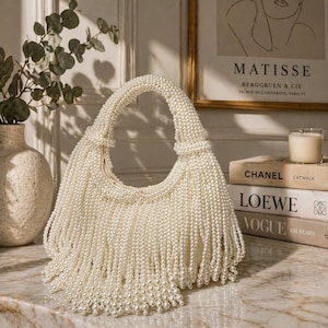 May include: Ivory-colored handbag adorned with numerous faux pearls, creating a fringed effect. The bag features a rounded shape with a handle also covered in pearls. The design is elegant and suitable for special occasions.