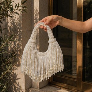 May include: A white beaded handbag with a rounded shape and a fringe of hanging beads. The handles are also beaded. The bag is held up against a neutral background with sunlight.