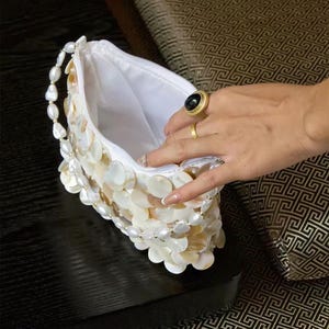 May include: A white clutch bag covered in overlapping iridescent shell discs, featuring a handle made of white oval beads. The bag has a white fabric interior lining and a zipper closure along the top edge, showcasing a textured, layered design.
