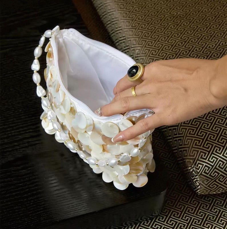 May include: A white clutch bag covered in overlapping iridescent shell discs, featuring a handle made of white oval beads. The bag has a white fabric interior lining and a zipper closure along the top edge, showcasing a textured, layered design.