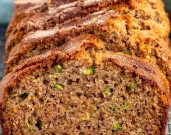 Puede incluir: Pan de calabacín en rodajas sobre una rejilla. El pan tiene una corteza dorada con azúcar en polvo. Se ven trozos verdes de calabacín en el interior húmedo y texturizado. Un delicioso producto horneado casero.