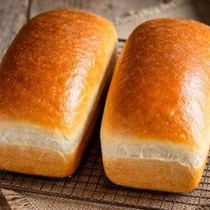 May include: Two loaves of golden-brown bread with a glossy crust sit on a wire rack. The bread has a rectangular shape with a light-colored bottom. The loaves are placed on a rustic wooden surface.