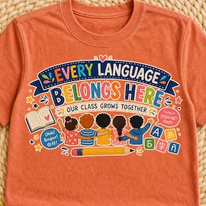 May include: A rust-colored t-shirt featuring a graphic of five children standing side-by-side with arms around each other. The text reads "EVERY LANGUAGE BELONGS HERE" and "OUR CLASS GROWS TOGETHER" alongside illustrations of a book, a pencil, and various language characters.