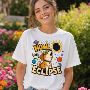 May include: A white t-shirt featuring a graphic print of a brown puppy wearing sunglasses and a collar, looking up at a solar eclipse. The text HOWL AT THE ECLIPSE is printed in yellow and black, surrounded by colourful planets and stars.