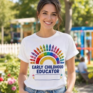 May include: White t-shirt featuring a rainbow design made of crayons and a ruler. The words "EARLY CHILDHOOD EDUCATOR" are printed below the rainbow, with a blank space. The shirt is worn by a person.