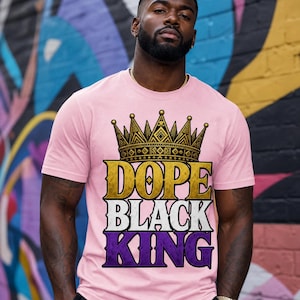 Op de afbeelding: Een lichtroze t-shirt met korte mouwen en een grafische print van een gouden kroon boven de tekst DOPE in goud, BLACK in wit en KING in paars. Het ontwerp is gecentreerd op de borst van het kledingstuk, dat een ronde hals heeft.