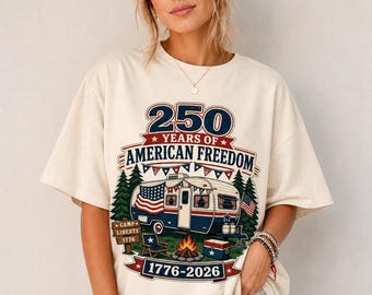 May include: An off-white t-shirt featuring a graphic of a vintage camper decorated with American flags and bunting. The text reads 250 YEARS OF AMERICAN FREEDOM, CAMP LIBERTY 1776, and 1776-2026. The design includes pine trees, a campfire, and a cooler.