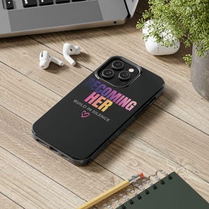Puede incluir: Funda de teléfono negra con el texto "BECOMING HER" en degradado rosa, morado y amarillo. La frase "BUILD IN SILENCE" y un corazón rosa están debajo. Sobre una superficie de madera con un portátil, auriculares y un cuaderno.