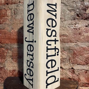 Puede incluir: Un letrero de madera de color claro con las palabras "Westfield" y "new jersey" impresas en negro. El letrero es rectangular y se alza sobre una estantería de madera oscura contra una pared de ladrillo.