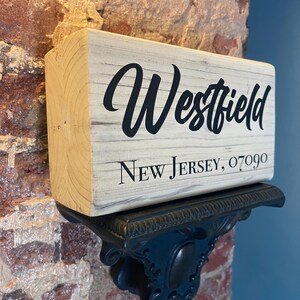Peut inclure: Panneau en bois avec le nom "Westfield" en écriture noire, et "New Jersey, 07090" en police serif. Le panneau est posé sur une étagère décorative en métal bleu foncé, contre un mur de briques.