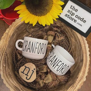 May include: Two white ceramic mugs with the name "Cranford" in black lettering are nestled in a wooden bowl with decorative rope and a sunflower. A wooden piece with "nj" is also in the bowl. A sign reads "the zip code store.com".