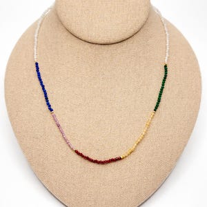 May include: A beaded necklace with a rainbow color scheme, displayed on a mannequin. The necklace features small, round beads in white, blue, pink, red, yellow, and green, separated by gold-colored spacers. The necklace is a jewelry item.