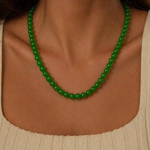 Puede incluir: Un collar de cuentas verdes. El collar está compuesto por cuentas redondas y pulidas en un tono verde vibrante. El collar se lleva alrededor del cuello. Las cuentas parecen ser de tamaño uniforme y están muy juntas.