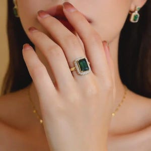 Puede incluir: Un anillo de oro con una piedra preciosa rectangular de color verde oscuro, rodeada de pequeñas piedras transparentes. El anillo está en una mano, con un pendiente a juego visible. La joya tiene un diseño clásico y elegante.