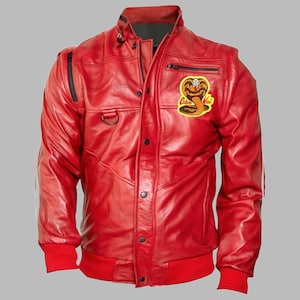 Puede incluir: Una chaqueta de cuero rojo brillante con cierre de cremallera frontal, tapeta con botones a presión y puños y dobladillo acanalados. En el pecho izquierdo, un parche bordado en amarillo y negro muestra un gráfico de cobra y el texto Cobra Kai.