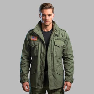 Könnte beinhalten: Eine olivgrüne Feldjacke mit Stehkragen, Frontreißverschluss und vier großen Taschen mit Knopfklappen. Auf der rechten Brust befindet sich ein rechteckiger Aufnäher der US-Flagge, auf der linken Brust der schwarz gestickte Schriftzug U.S. ARMY.