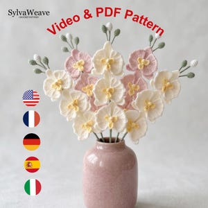 Può includere: Un bouquet di orchidee all'uncinetto in un vaso rosa. I fiori sono bianchi e rosa con centri gialli. L'immagine include il testo "Video & PDF Pattern" e il logo SylvaWeave.