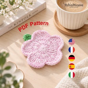Może przedstawiać: Jasnoróżowa szydełkowana podstawka w kształcie kwiatka z małym zielonym listkiem. Tekst PDF Pattern jest wyświetlany na czerwono. Obraz zawiera biały kubek do kawy, książkę zatytułowaną The Kinfolk Home oraz pięć okrągłych ikon flag.