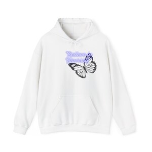Puede incluir: Una sudadera con capucha blanca que presenta un gráfico de mariposa negra y el texto "Believe in Yourself" en letra cursiva de color morado claro. La prenda incluye un bolsillo tipo canguro frontal, mangas largas y capucha con cordones blancos.