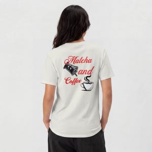 Puede incluir: Una camiseta de manga corta color crema con un gráfico en la parte posterior. El diseño incluye el texto rojo "Matcha and Coffee" con una ilustración en blanco y negro de una bebida helada y una taza de café humeante bajo el texto.