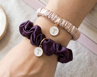 May include: Two fabric hair scrunchies worn on the wrist, one in a deep purple and one in a pale blush pink. Each scrunchie features a small, round, white enamel charm with the letter T printed in gold, attached by a thin gold-toned metal ring.