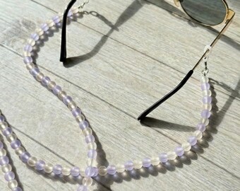 May include: Gold-framed sunglasses with dark lenses rest on a light wood surface, connected to a beaded eyeglass chain. The chain features alternating clear and lavender-colored beads, adding a touch of color and style to the accessory.