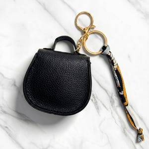 May include: A black leatherette mini handbag keychain with a gold-tone clasp and key ring. The keychain includes a patterned scarf-like strap. The bag has a top handle and a textured surface.