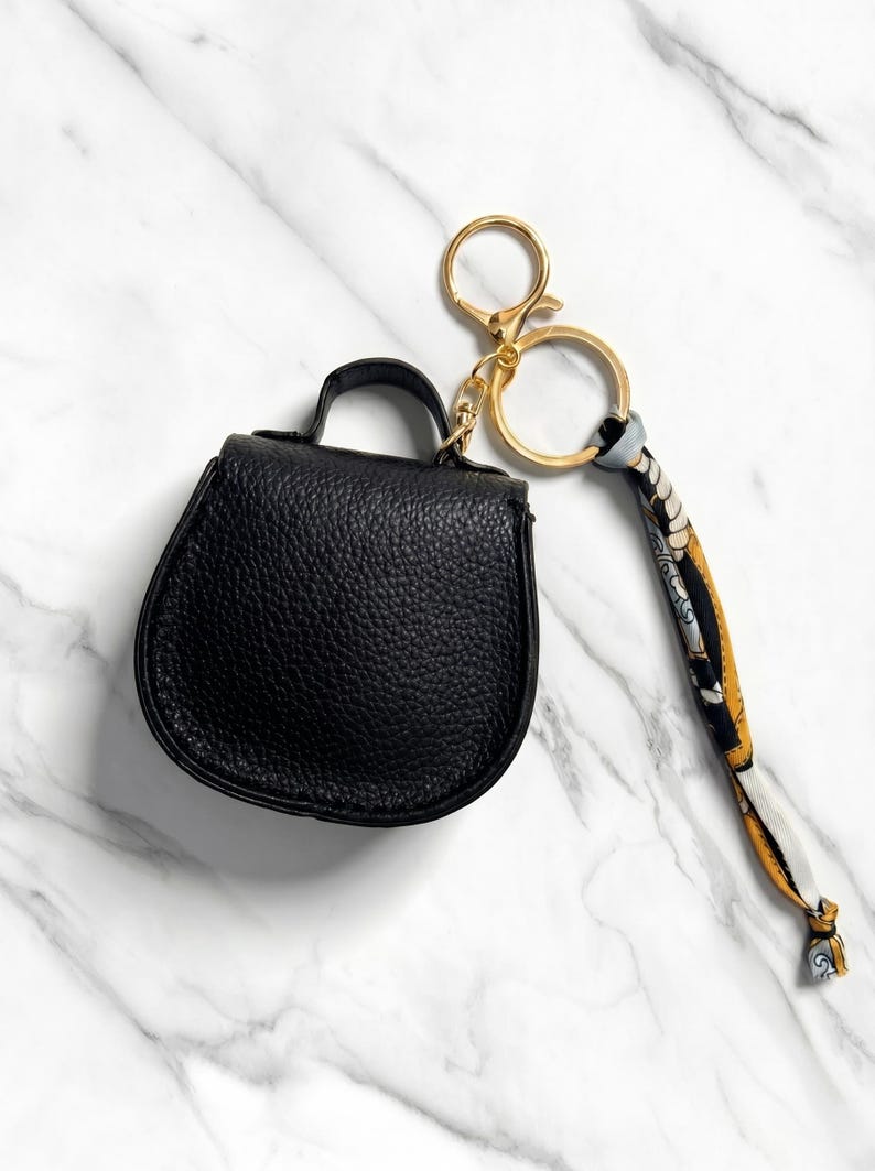 May include: A black leatherette mini handbag keychain with a gold-tone clasp and key ring. The keychain includes a patterned scarf-like strap. The bag has a top handle and a textured surface.