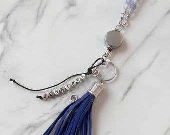 May include: A personalized lanyard with the name "JENNIFER" spelled out in silver letter beads. The lanyard features a blue tassel, a retractable reel, and a beaded chain with purple and clear beads. The background is a white marble surface.