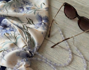 May include: A set including a floral scarf and an eyeglass chain. The scarf features a white background with blue floral patterns and a navy blue border. The eyeglass chain is made of light purple beads and is attached to a pair of sunglasses.