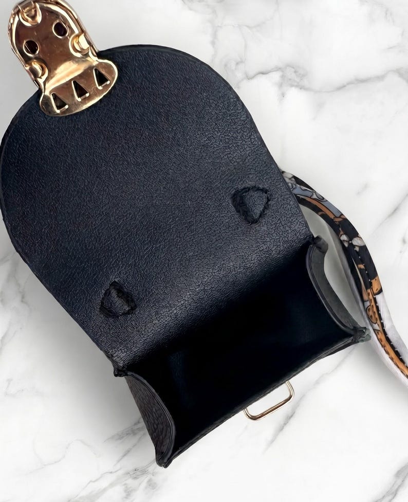 May include: Black leather mini bag with a gold-tone clasp and hardware. The bag has a structured shape and a patterned strap. The interior is black. The bag is open, revealing the inside.