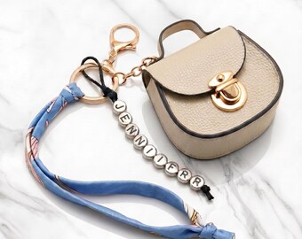 May include: A beige miniature handbag keychain with a gold-tone clasp and a top handle. Attached is a blue fabric strap and a black cord featuring white letter beads spelling out JENNOOFRR, connected by a gold-tone metal ring and lobster claw.