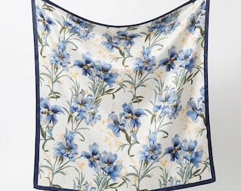 May include: Square scarf with a floral pattern. The scarf features a repeating design of blue flowers with green stems and leaves on a white background. The edges are trimmed with a dark blue border.