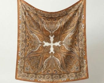 May include: A square scarf with a paisley pattern in shades of brown, tan, and cream. The scarf has a border with a repeating design and is hanging against a white background. The scarf is made of a soft, flowing material.