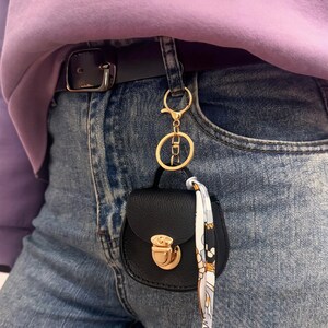 May include: A small, black handbag keychain with a gold clasp and hardware is attached to a belt loop. A patterned scarf hangs from the keychain. The person is wearing blue jeans and a purple sweatshirt.