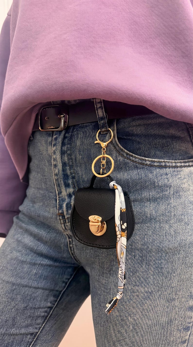 May include: A small, black handbag keychain with a gold clasp and hardware is attached to a belt loop. A patterned scarf hangs from the keychain. The person is wearing blue jeans and a purple sweatshirt.