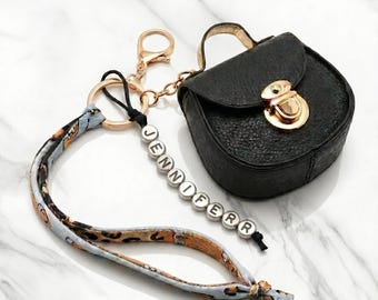May include: A black leatherette purse keychain with a gold clasp and hardware. The keychain includes a patterned fabric strap and a beaded name tag that reads "JENNIFER".
