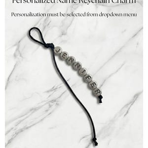 May include: A personalized name keychain charm with the name "JENNIFER" spelled out in silver letter beads. The keychain has a black cord and the text "Custom text up to 15 characters".
