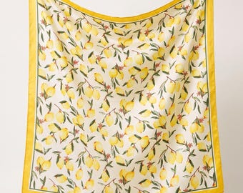 May include: A square scarf featuring a repeating pattern of yellow lemons, green leaves, and small red blossoms on a cream background. The scarf is framed by a solid yellow border with a thin dark green line accentuating the lemon print design.