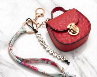 May include: A red textured mini bag keychain with a gold-tone clasp, featuring a light blue fabric ribbon with a floral pattern and a black cord string of white beads spelling the name JENNIFER, all attached to a gold-tone metal ring.