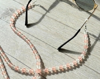 May include: Gold-framed sunglasses with dark lenses rest on a light wooden surface, accompanied by a beaded eyeglass chain. The chain features alternating pink and clear beads, adding a touch of color and style to the accessory.